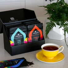Load image into Gallery viewer, Discovery Box with luxury organic loose leaf teas and collagen peptides on kitchen counter, Pure Detox and Cronulla Chai displayed at front of box with green plant and yellow tea cup and saucer with Elouera Earl French Earl Grey tea in cup and tea pouch with pretty tea leaves spilling out on to counter. Satin finish black box with 9 pouches of loose leaf tea and 1 pouch of collagen peptides powder. Gift Pack. Loose leaf tea gift pack. 10 pouches standing in 2 rows of 5 in box.