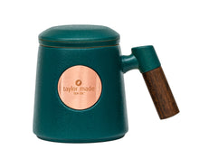 Load image into Gallery viewer, Front view of textured matt teal coloured ceramic three-piece tea cup with modern design and wooden handle. Circular rose gold metal panel on front with etched Taylor Made Tea Co. logo.  Matching infuser is inserted into the cup with matching teal rim and matching teal lid sits upon cup and infuser.
