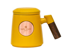 Load image into Gallery viewer, Front view of textured matt yellow ceramic three-piece tea cup with modern design and wooden handle. Circular rose gold metal panel on front with etched Taylor Made Tea Co. logo.  Matching infuser is inserted into the cup with matching yellow rim and matching yellow lid sits upon cup and infuser.
