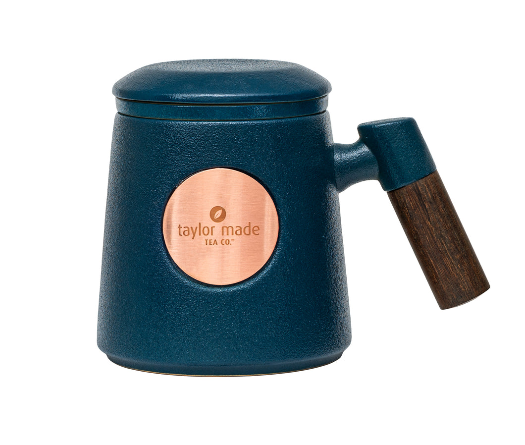 Front view of textured matt midnight blue ceramic three-piece tea cup with modern design and wooden handle. Circular rose gold metal panel on front with etched Taylor Made Tea Co. logo.  Matching infuser is inserted into the cup with matching blue rim and matching blue lid sits upon cup and infuser.