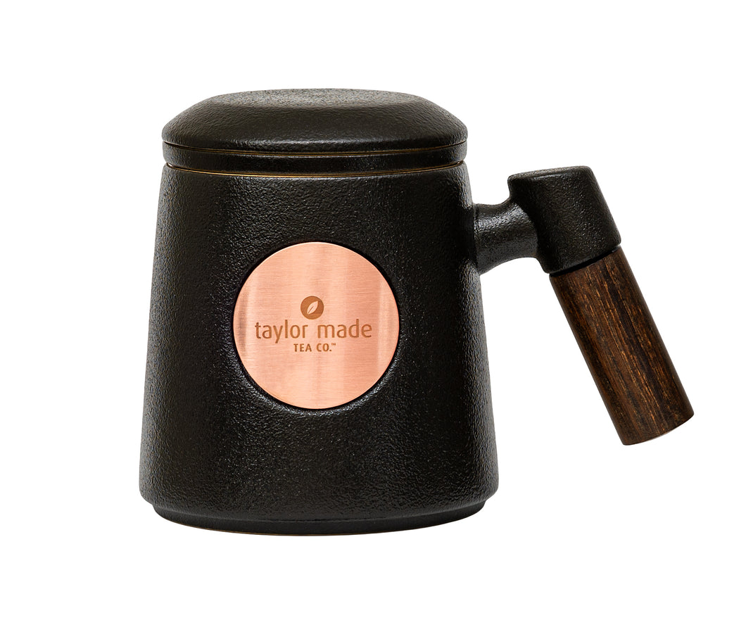 Front view of textured matt black ceramic three-piece tea cup with modern design and wooden handle. Circular rose gold metal panel on front with etched Taylor Made Tea Co. logo.  Matching infuser is inserted into the cup with matching black rim and matching black lid sits upon cup and infuser.