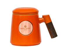Load image into Gallery viewer, Front view of textured matt bright orange ceramic three-piece tea cup with modern design and wooden handle. Circular rose gold metal panel on front with etched Taylor Made Tea Co. logo.  Matching infuser is inserted into the cup with matching orange rim and matching orange lid sits upon cup and infuser.
