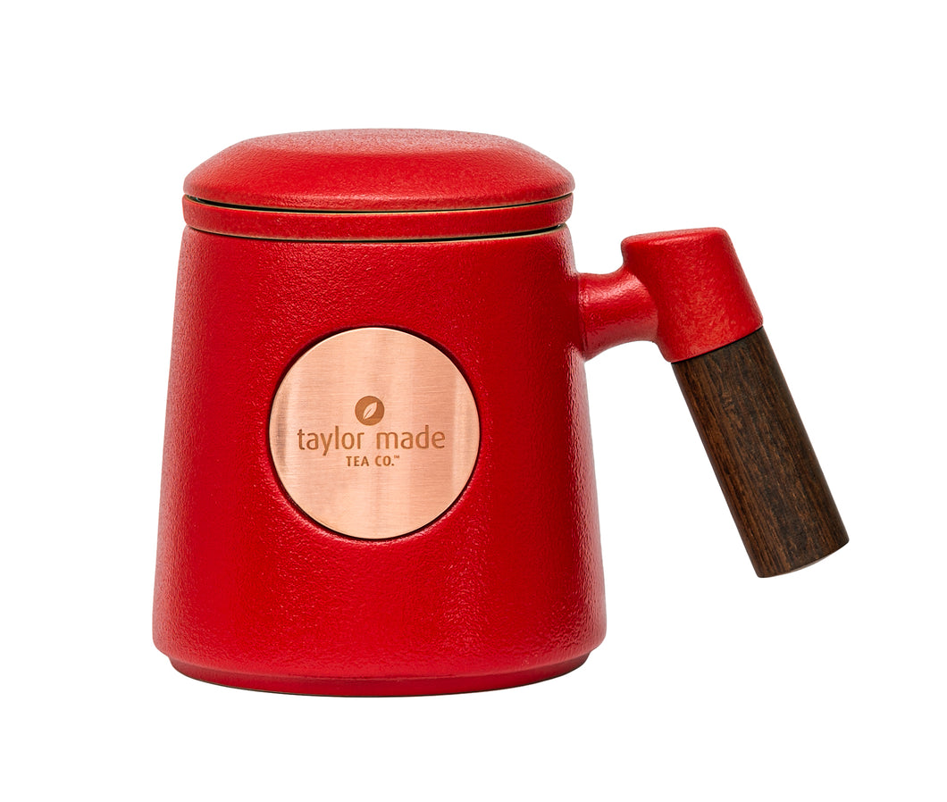 Front view of textured matt vivid red ceramic three-piece tea cup with modern design and wooden handle. Circular rose gold metal panel on front with etched Taylor Made Tea Co. logo.  Matching infuser is inserted into the cup with matching red rim and matching red lid sits upon cup and infuser.