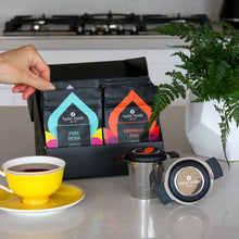 Load image into Gallery viewer, Discovery Box with luxury organic loose leaf teas and collagen peptides on kitchen counter, Pure Detox and Cronulla Chai displayed at front of box with green plant and yellow tea cup and saucer with 2 black signature infusers tea in cup.  Satin finish black box with 9 pouches of loose leaf tea and 1 pouch of collagen peptides powder. Gift Pack. Loose leaf tea gift pack. 10 pouches standing in 2 rows of 5 in box. 
